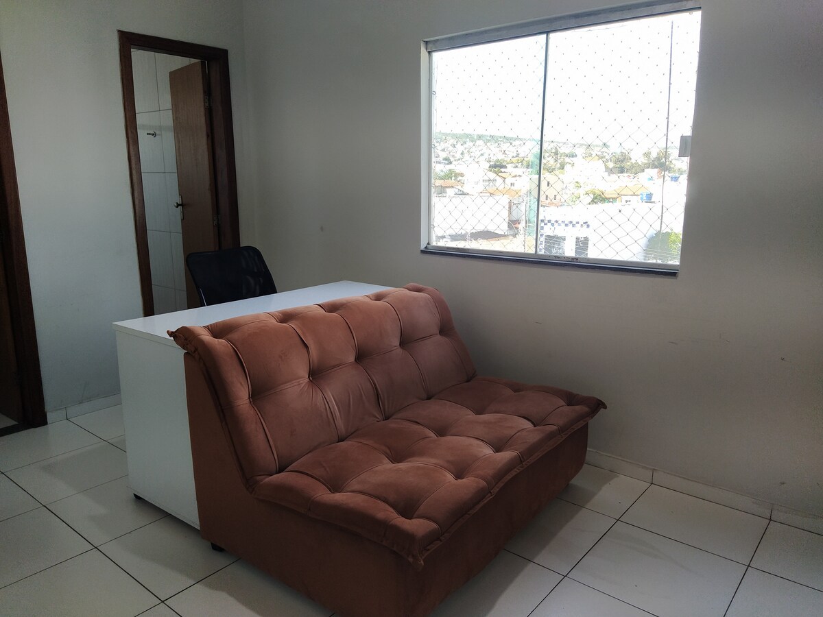 A comfortable sofa is positioned in a well-lit room, accompanied by a white desk and an office chair. A window allows natural light to fill the space, while a grid pattern on the window adds texture to the view.