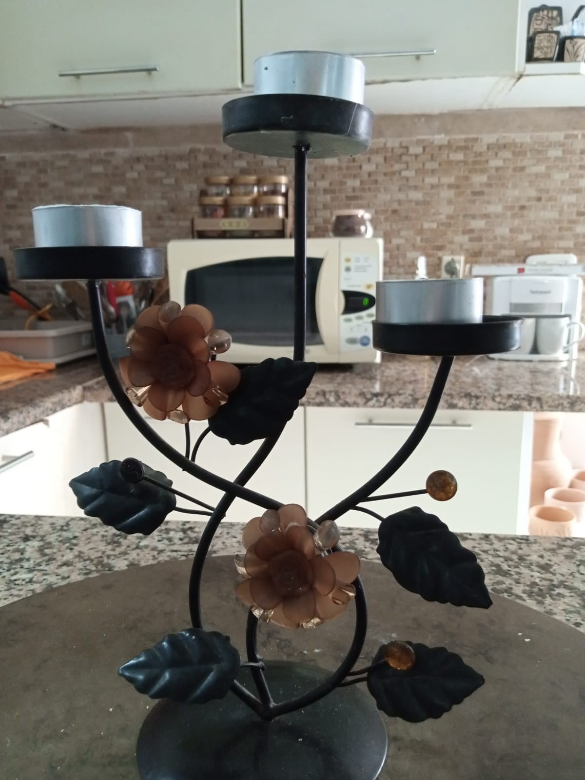 A decorative candle holder is presented on a stone surface, featuring a swirling design of dark leaves and brown flowers. The holder accommodates three candles and is set against a backdrop of a modern kitchen, with a glimpse of appliances and kitchenware in soft focus.