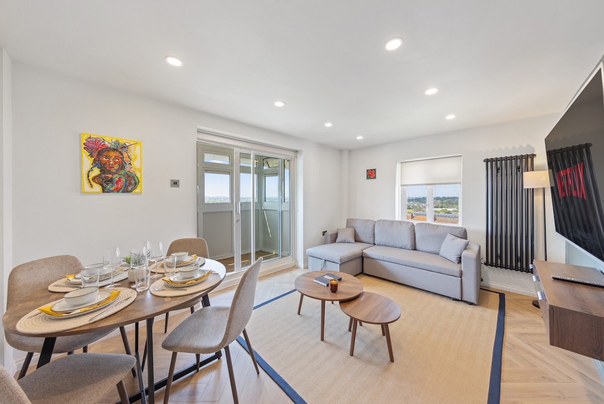 A modern living room is showcased, featuring a comfortable grey sofa and a round dining table set for six. Large windows allow natural light to illuminate the space. A vibrant piece of artwork adds a touch of color to the neutral decor.
