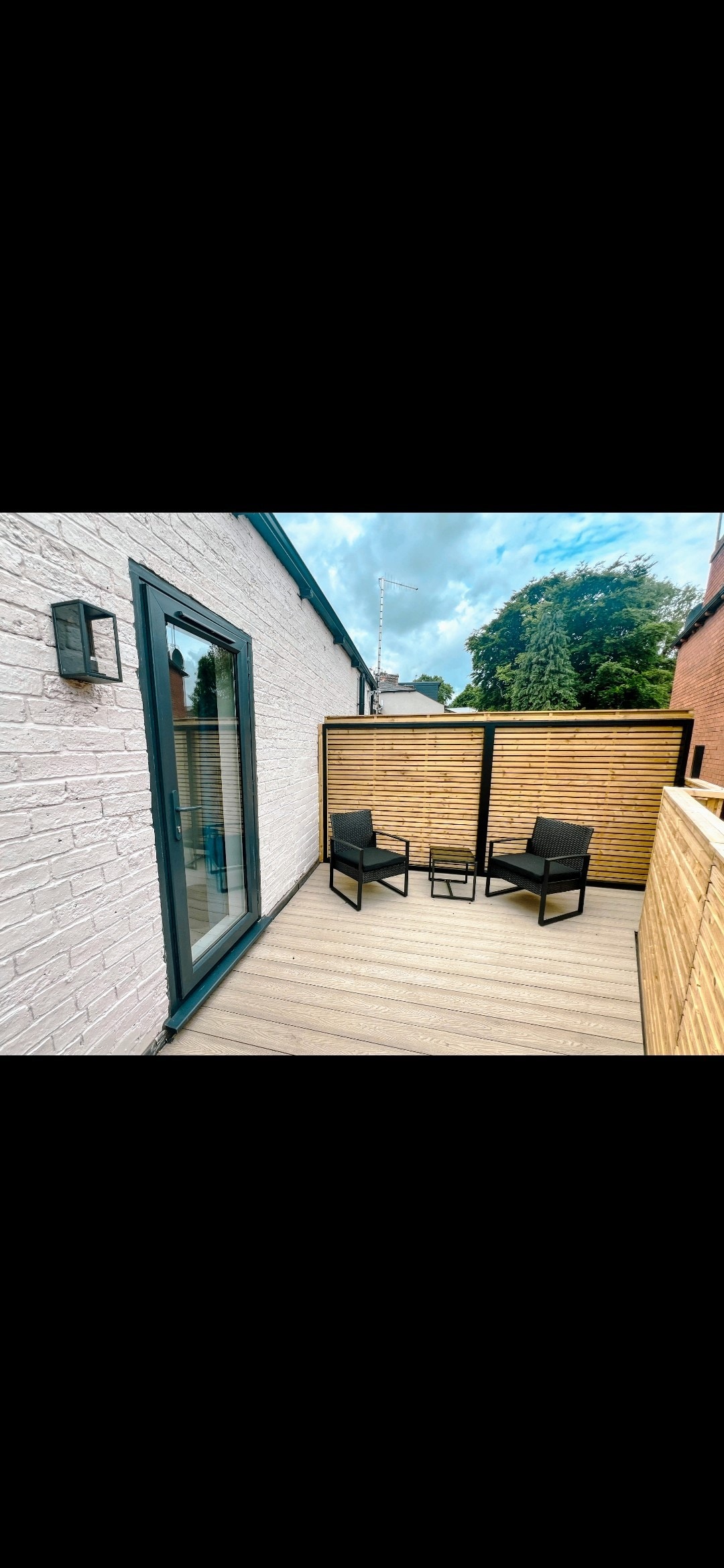 A private decked roof terrace is depicted, featuring two black chairs and a small table between them. Wooden slats provide privacy, while a glass door provides access to the interior. Lush greenery is visible in the background, enhancing the calming outdoor space.