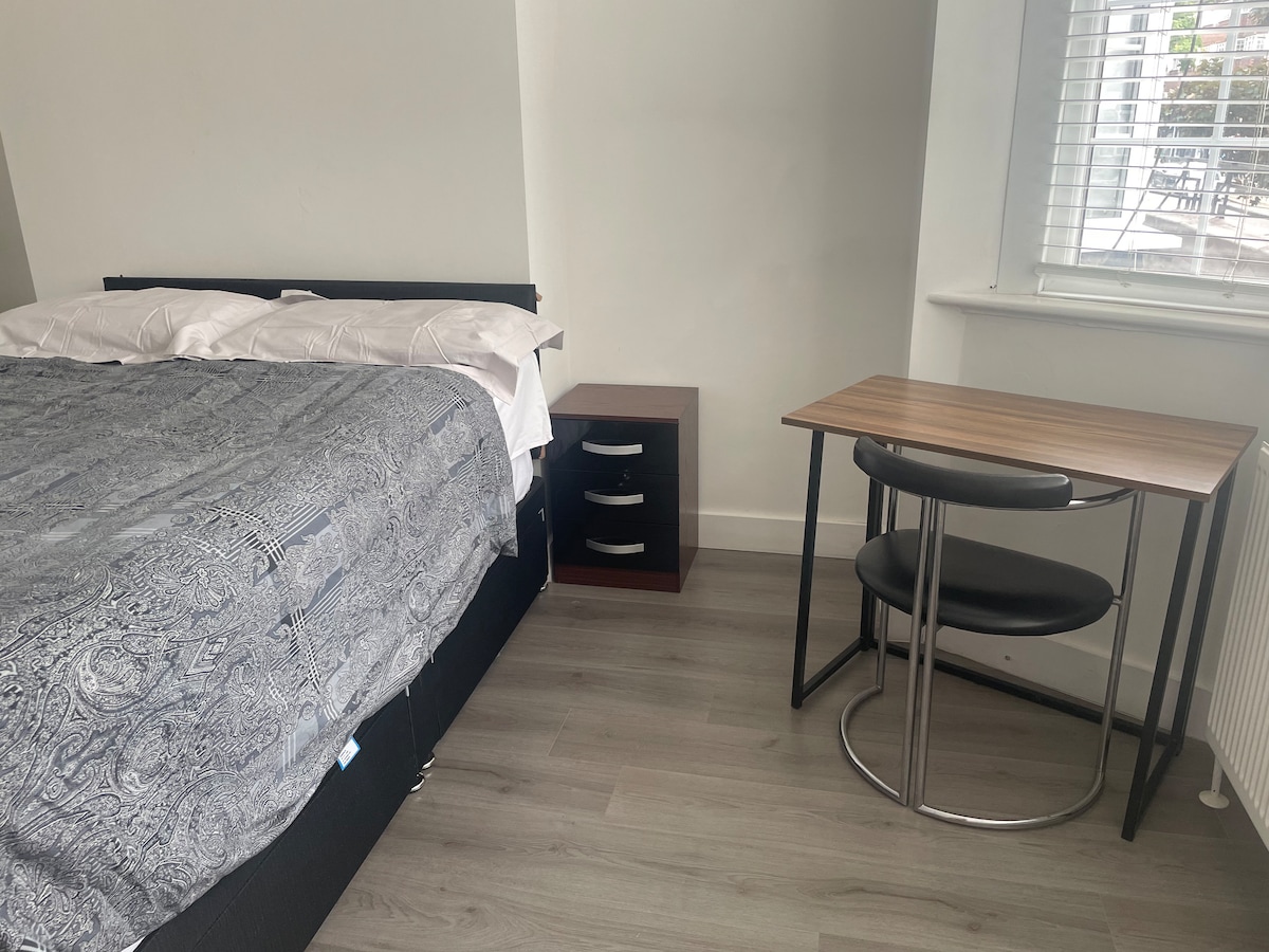 A double bed with a decorative gray bedspread is positioned against a wall. Next to the bed, a small nightstand with drawers is visible. A round table with black chairs is situated nearby, enhancing the room's functionality. Natural light filters through a partially opened window.