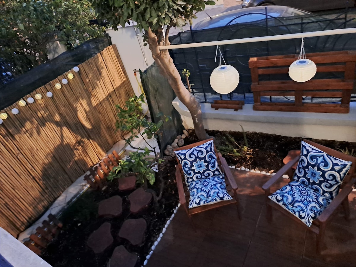 An outdoor terrace is presented with two wooden chairs adorned with decorative blue cushions. A small garden area is visible, featuring a tree and various plants. Soft lighting is provided by hanging lanterns, creating a calm and inviting space.