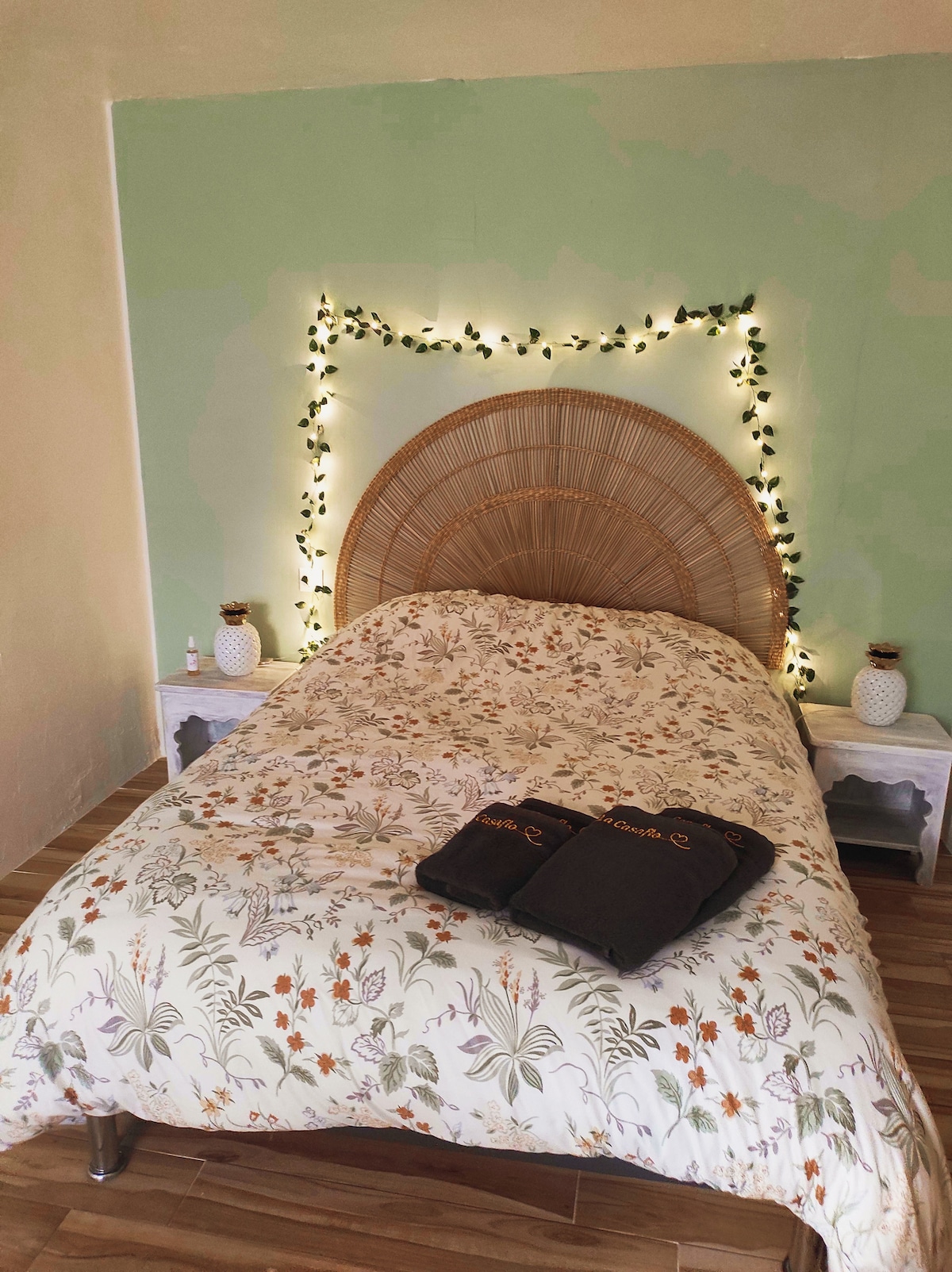 A cozy sleeping area features a spacious bed adorned with a floral-patterned comforter. A decorative rattan headboard is highlighted by soft, ambient lighting. Two small bedside tables are positioned on either side, accompanied by decorative items, creating a serene environment.