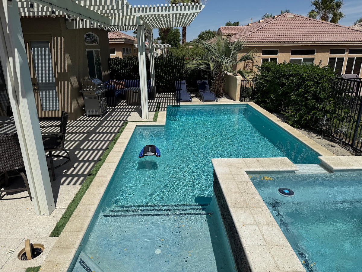 A serene backyard area features a sparkling swimming pool surrounded by paved surfaces. Lounge chairs are positioned nearby, offering a space for relaxation under a shaded pergola. Lush greenery lines the boundary, contributing to a private atmosphere.