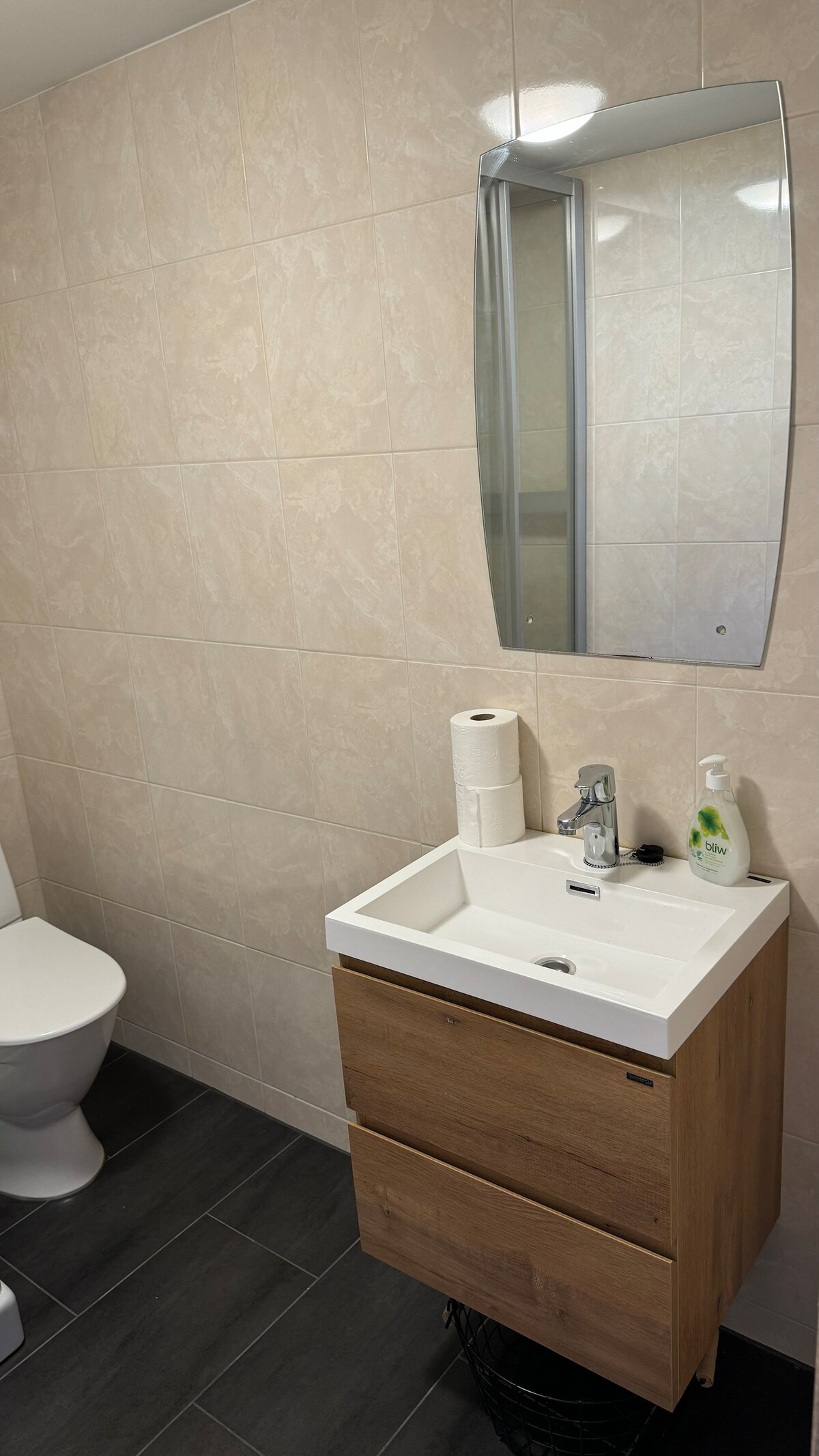 The bathroom features a modern design with light-colored tile walls and dark floor tiles. A compact wooden vanity with two drawers is equipped with a sink and a faucet. A wall-mounted mirror reflects the space, while toiletries are neatly arranged on the countertop.