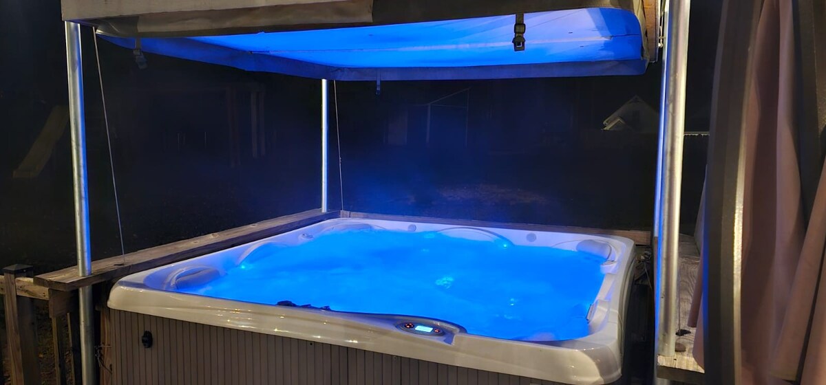 A hot tub sits on a deck, softly illuminated by blue lighting, creating a serene ambiance. The surrounding area features a partial canopy overhead, providing a sheltered space for relaxation. The misty atmosphere enhances the tranquil setting, ideal for unwinding under the night sky.