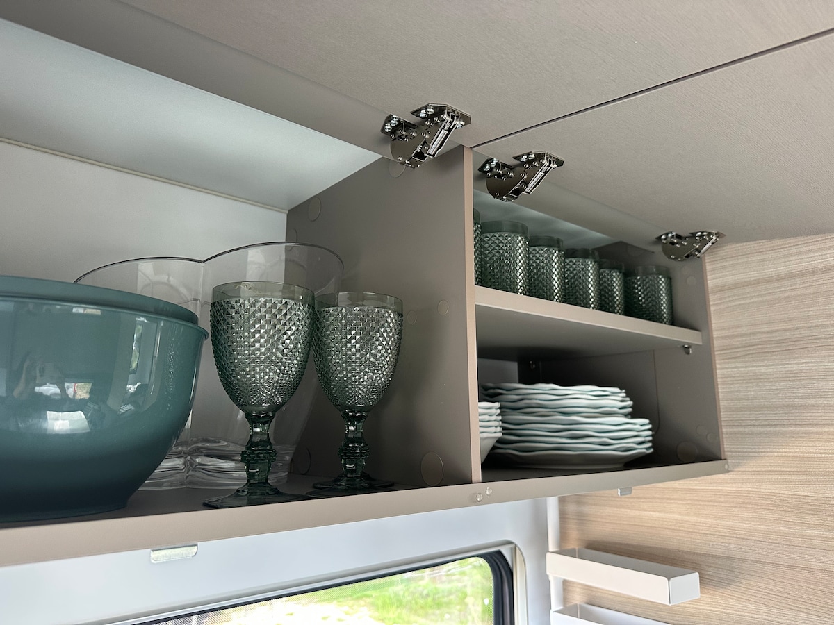 A well-organized cabinet interior displays a variety of glassware and tableware. Sparkling green goblets are stacked beside clear drinking glasses. A large blue bowl rests on the upper shelf, while neatly arranged white plates line the lower shelf, providing convenient dining essentials.