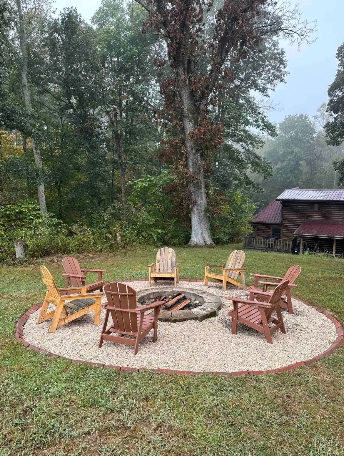 A fire pit surrounded by six wooden chairs is situated on a gravel patch, set amongst lush greenery. Tall trees frame the scene, while a rustic cabin with a metal roof is visible in the background, contributing to the serene atmosphere of the outdoor space.
