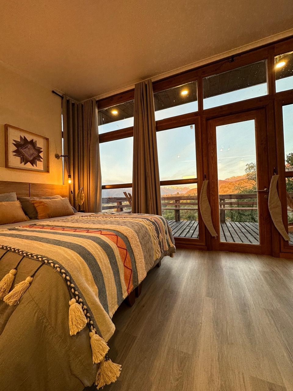 A cozy bedroom is highlighted by large windows showcasing scenic views of the surrounding landscape. A spacious bed is adorned with a textured blanket featuring earthy tones and decorative fringes. Soft lighting creates a serene ambiance, complemented by natural wood accents throughout the room.