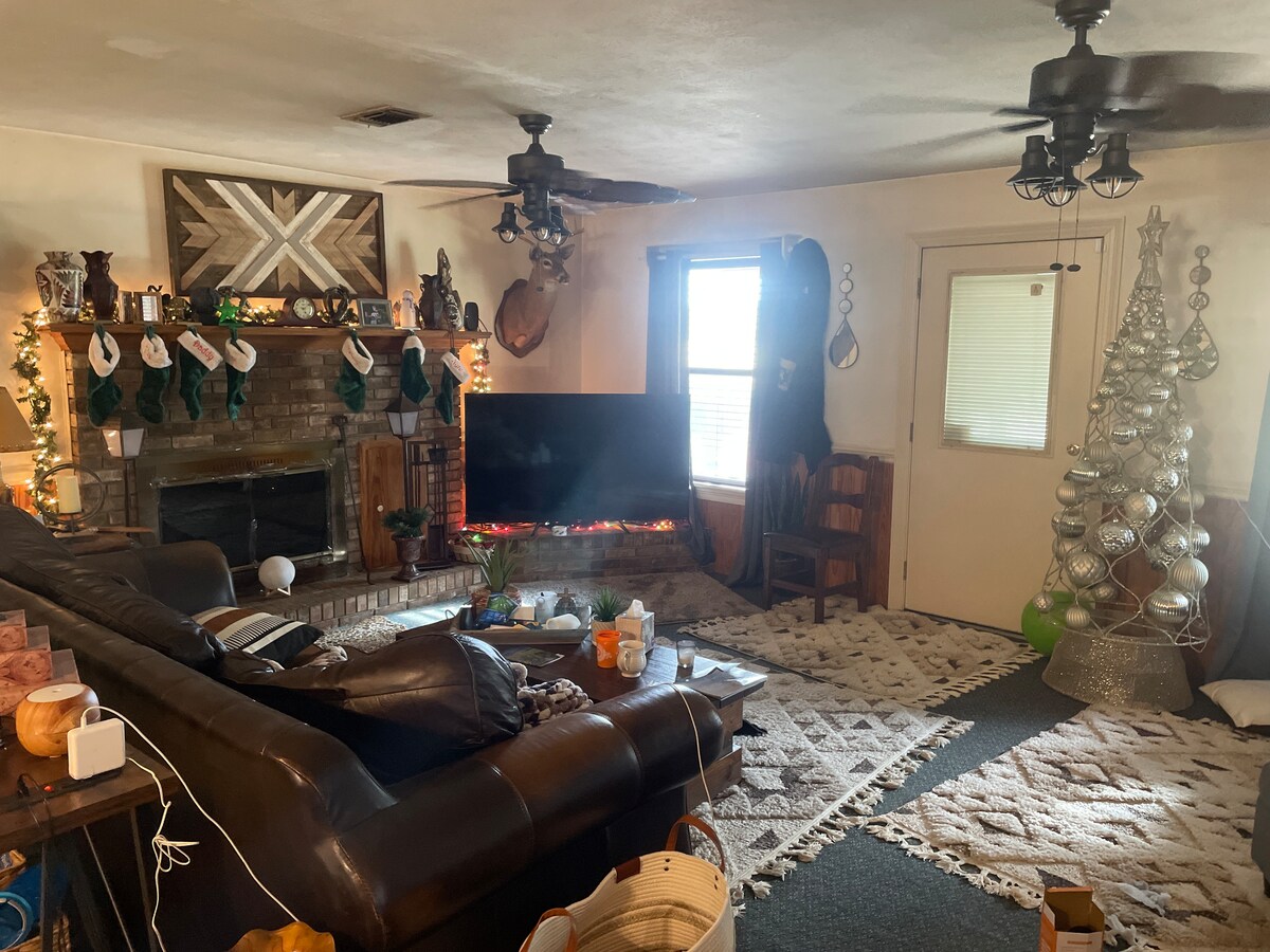 A cozy living area features a large dark sectional couch facing a wall-mounted television. The fireplace is adorned with stockings and decorative items. A silver Christmas tree stands nearby, contrasting with the warm tones of the room. Natural light filters in through the window.