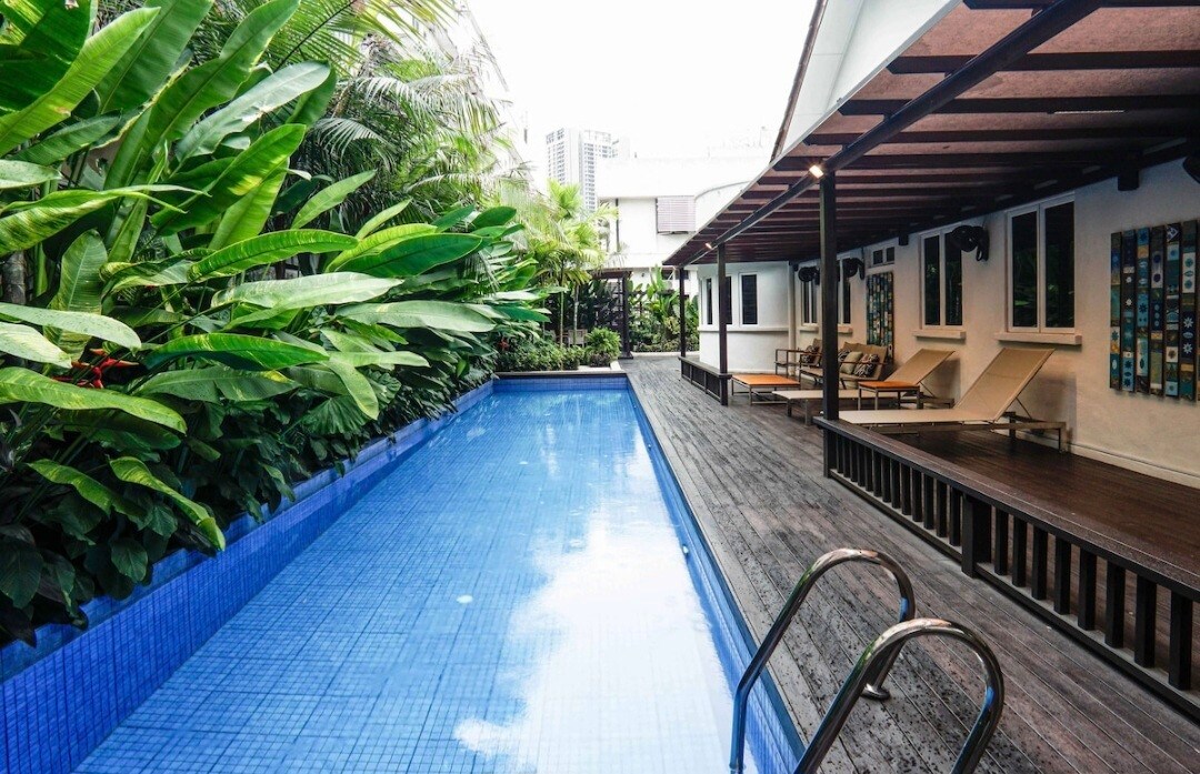 A tranquil swimming pool is framed by lush green plants, providing a serene backdrop. Sun loungers are positioned on the adjacent wooden deck, inviting relaxation. The setting showcases a blend of nature and modern design, emphasizing a peaceful retreat.