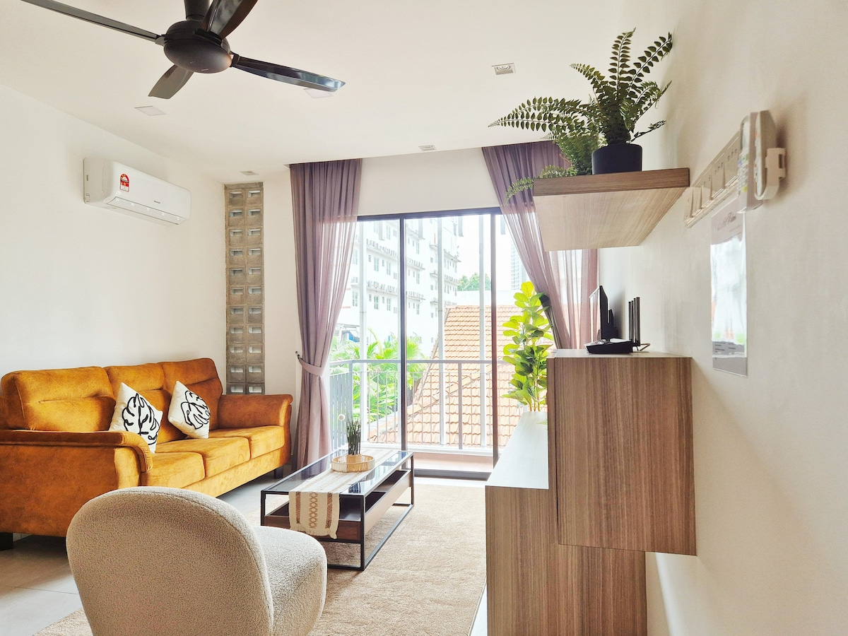 A bright living area features a comfortable orange sofa and a stylish light-colored armchair. Large windows provide natural light and a view of greenery outside, while a wooden shelf displays decorative elements. A coffee table with a glass top is positioned in front of the seating arrangement.