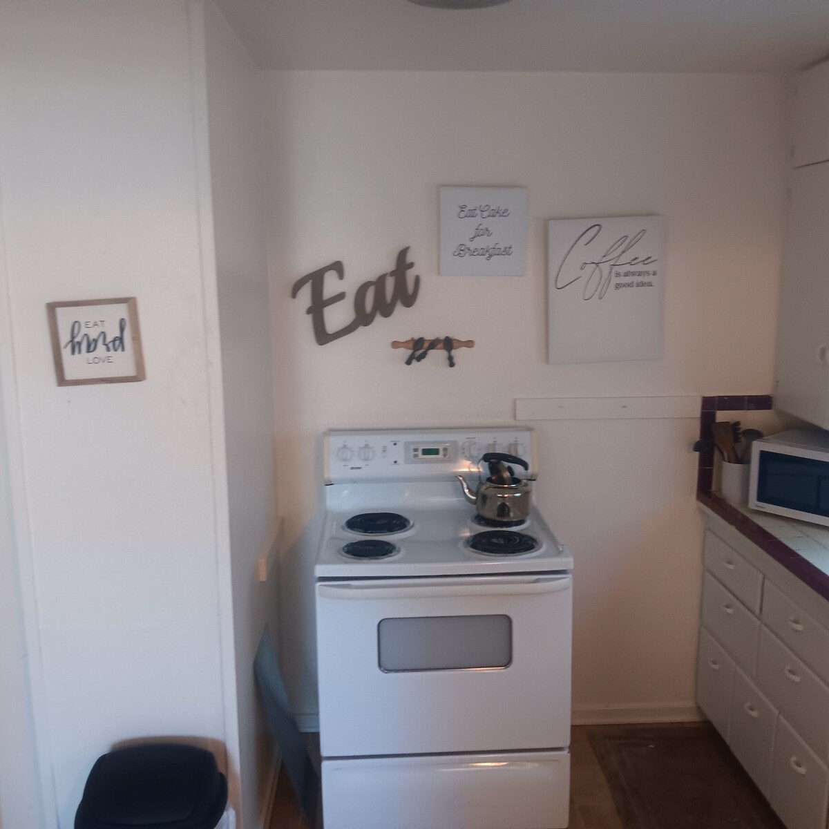 A compact kitchen is presented, featuring a white electric stove with four burners and an oven below. Wall art with phrases about eating and coffee is displayed, along with a small microwave and a pot on the stove, contributing to a functional cooking space.