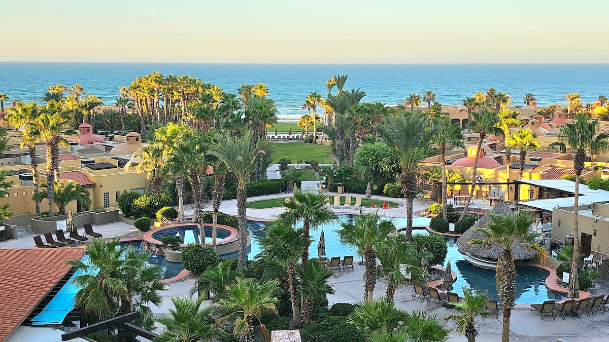 A view of the resort’s grounds showcases several palm trees surrounding multiple pools. Lounge chairs are arranged near the water, and a sandy beach extends to the horizon. The ocean is visible in the background, adding a tranquil ambiance.