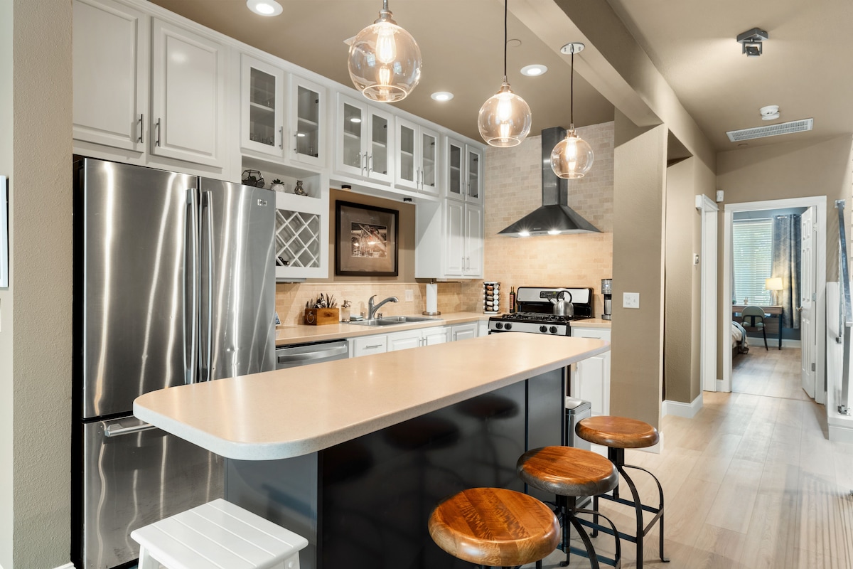 The kitchen showcases modern cabinetry with glass display sections and stainless steel appliances. A central island features three wooden stools. Pendant lighting hangs above, illuminating the space. The open design leads to a hallway that connects to other areas of the townhouse.