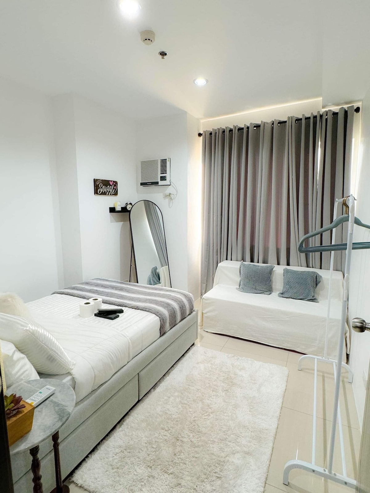 A compact room features a neatly made spacious bed with soft linens. A small white sofa with decorative cushions is positioned opposite. A full-length mirror reflects the natural light from the window, which is dressed in stylish gray curtains. A small round table adds to the decor.