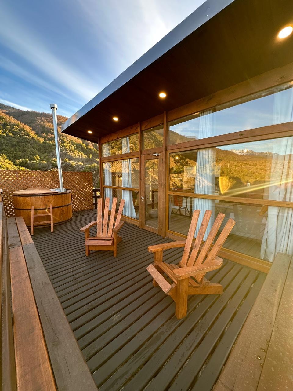 A spacious outdoor deck features two wooden adirondack chairs positioned for relaxation. A hot tub is visible nearby, surrounded by wooden lattice for privacy. Large glass doors provide access to the interior, with scenic views of the surrounding natural landscape.