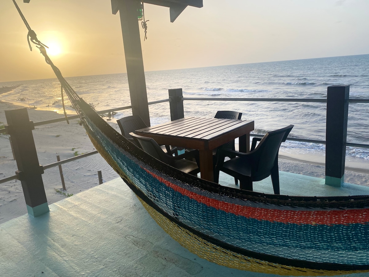A hammock with colorful stripes is hung under a shaded area, accompanied by a wooden table and several black chairs. The ocean's waves are visible in the background, along with a sunset casting a warm glow over the scene.
