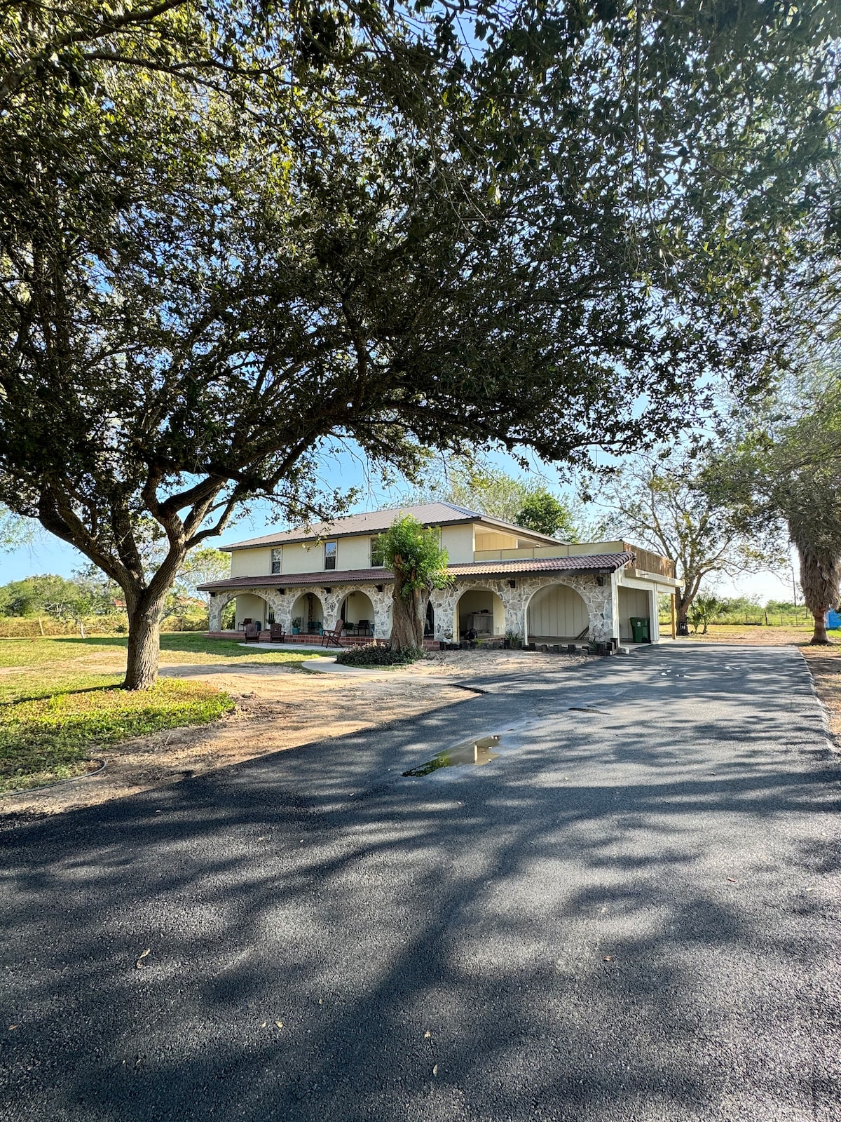 The exterior of a two-story home is displayed, surrounded by large trees that provide shade. A paved driveway leads up to the entrance, while an expansive grassy area is visible in the background under a clear blue sky.