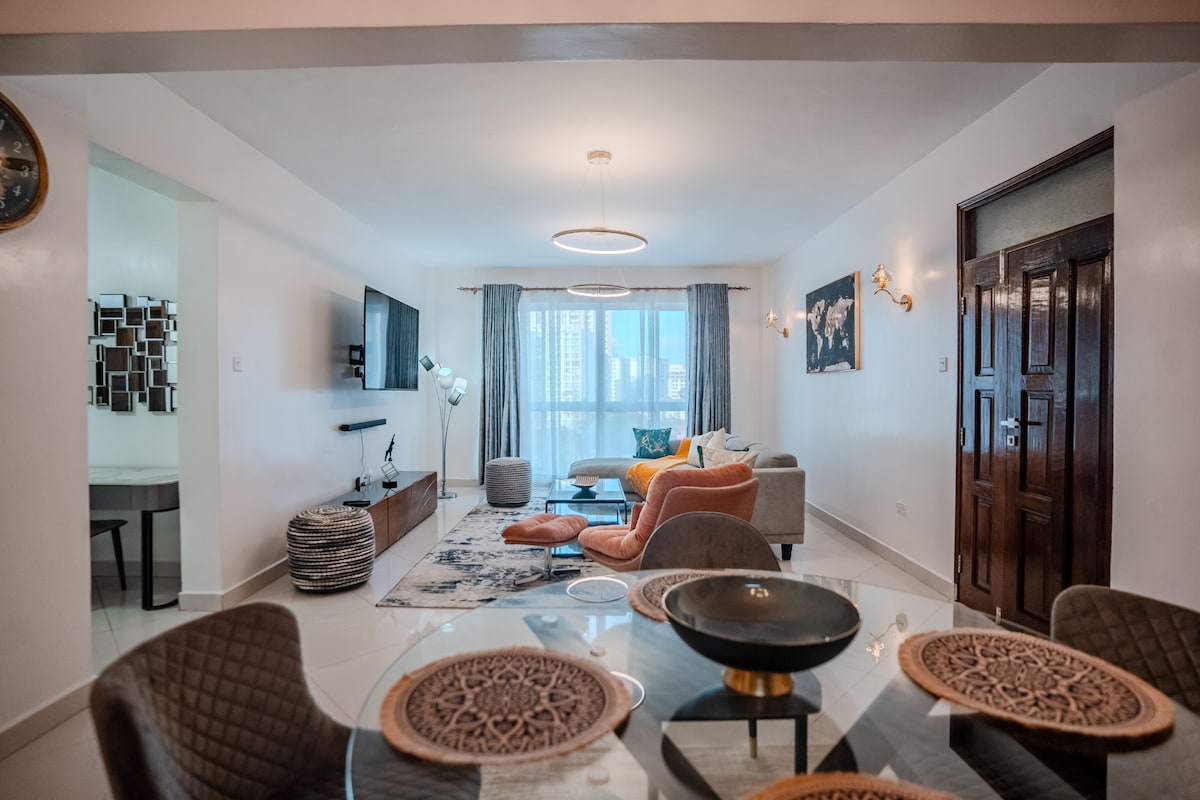 A modern living area is showcased, featuring a large sofa, two accent chairs, and a coffee table. A round dining table with woven placemats is positioned nearby. Natural light enters through the large window, illuminating the space, which is decorated with wall sconces and artwork.