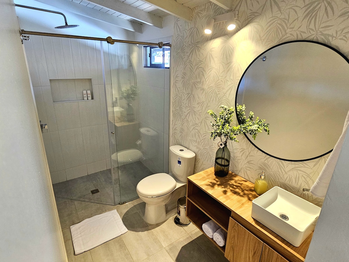 The bathroom features a glass shower enclosure and a round mirror above a wooden vanity. A vase with greenery adds a touch of nature, while white towels are neatly arranged beside the sink. Natural light enters through a small window.