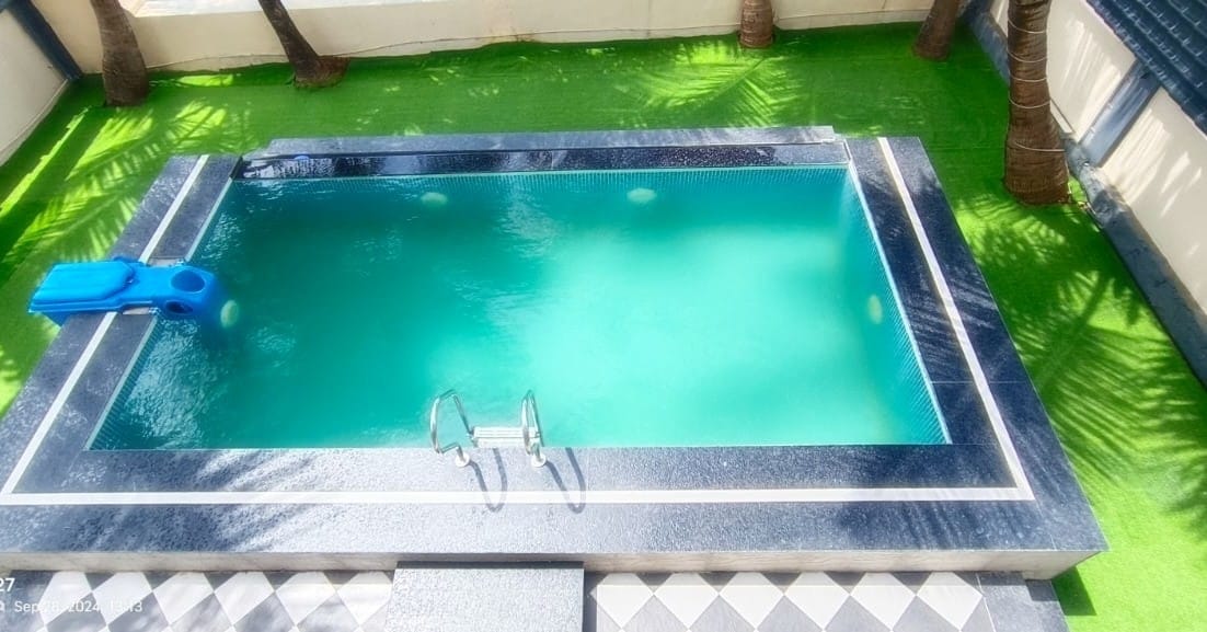 A serene private swimming pool is showcased, surrounded by manicured green grass. Palm trees are visible along the edges, providing a natural touch to the space. The pool features steps leading in and is accented by a smooth dark stone border.