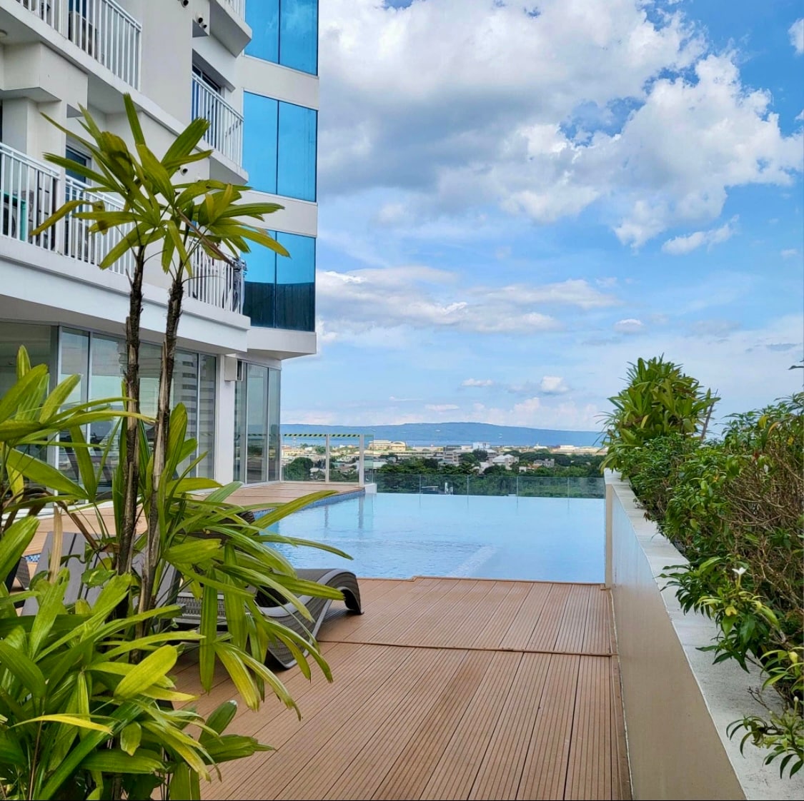 An outdoor infinity pool is visible, surrounded by wooden decking and lush greenery. The serene water reflects the sky, with distant hills and buildings in the background. Sun loungers are positioned nearby, inviting relaxation amid a tranquil setting.