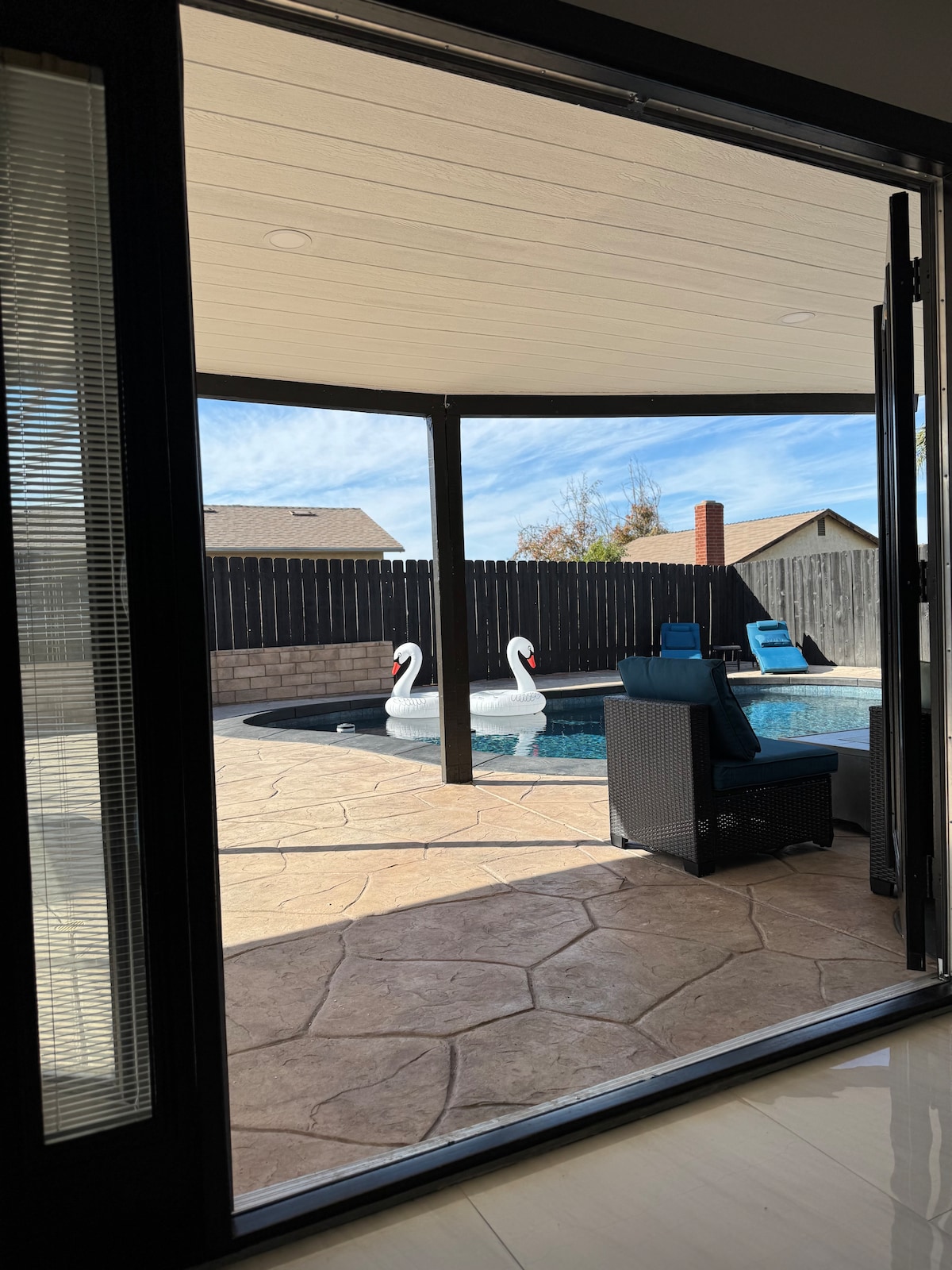 A view of the outdoor area highlights a swimming pool surrounded by a stone patio. Two swan-shaped floatation devices rest on the water, while a modern chair with blue cushions is positioned nearby. Glass doors provide a seamless transition from indoors to the outdoor space.