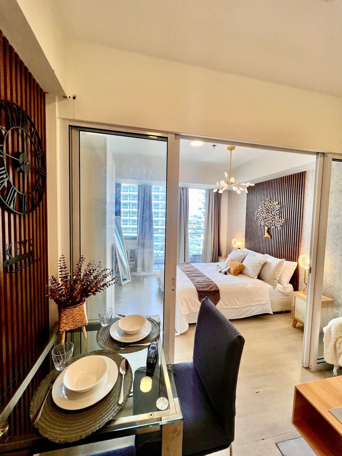 A dining area with a table set for four is visible, featuring dinnerware and glassware. The spacious bedroom with a large window is seen in the background, showcasing a queen-sized bed with neutral bedding against a textured wall.