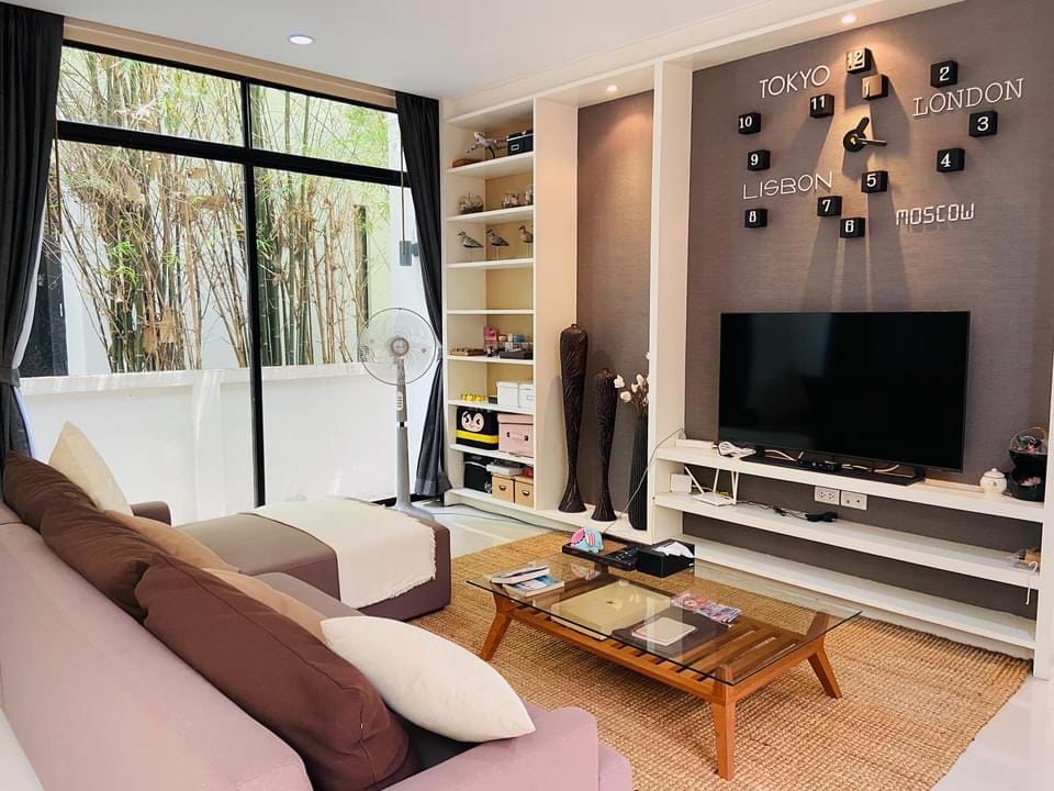 A modern living area features a large window allowing natural light to illuminate the space. Two sofas in neutral tones are positioned around a glass coffee table. A wall-mounted TV and a decorative arrangement of city names enhance the contemporary design.