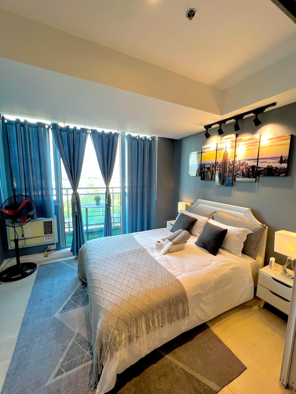 The bedroom showcases a spacious queen-sized bed, dressed with crisp white linens and plush pillows. Floor-to-ceiling curtains frame a window offering natural light. A decorative wall art piece reflects a city skyline, while bedside tables provide convenience. A ceiling lamp adds soft illumination.