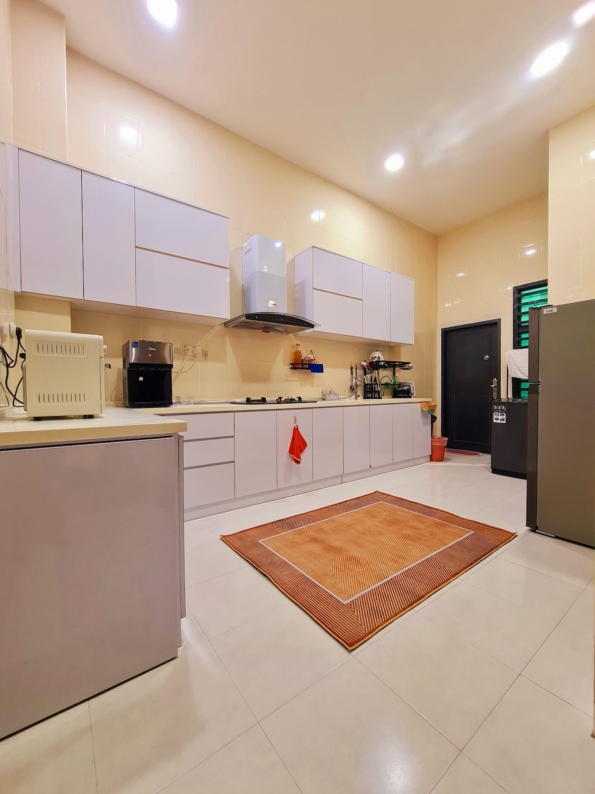 The kitchen features modern cabinetry in light tones, complemented by a neutral-colored rug on the tiled floor. Essential appliances, including a refrigerator and coffee maker, are visible, while a window allows natural light to enhance the space.