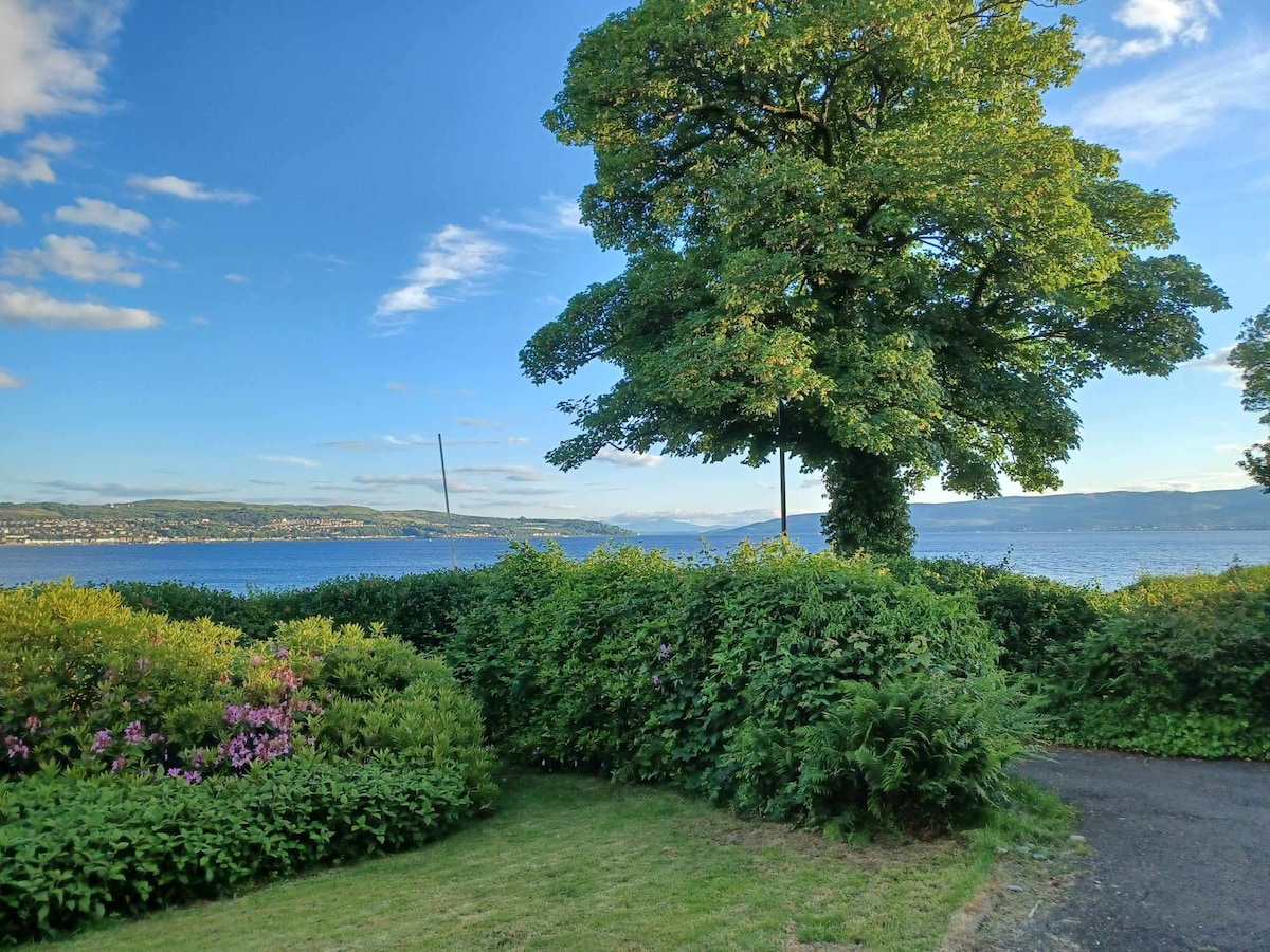 A view of the serene waterfront is framed by lush greenery, including a large tree and neatly trimmed hedges. The scene captures a vibrant blue sky with a few scattered clouds, while the shimmering water reflects the surrounding landscape.