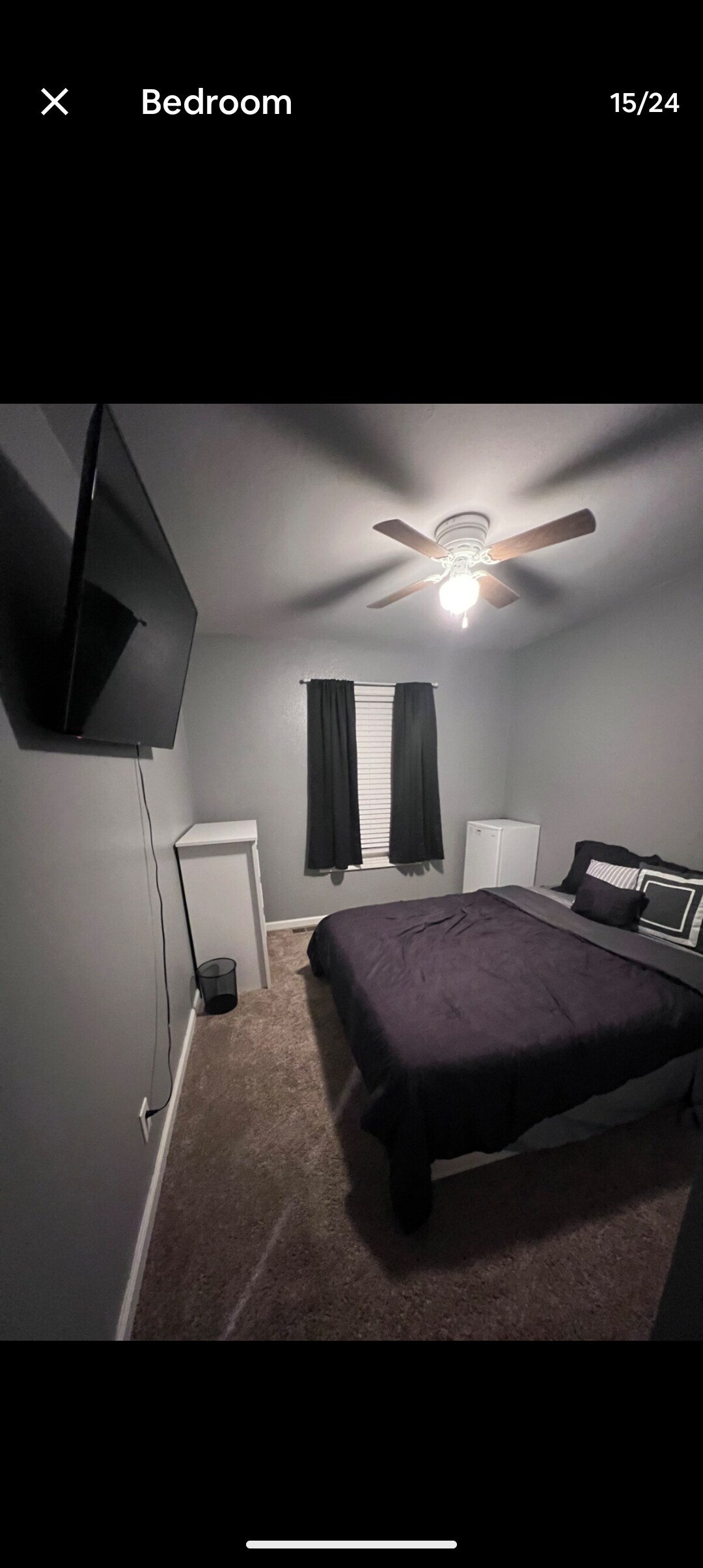 A cozy bedroom features a spacious bed dressed in dark bedding. A ceiling fan provides ventilation, while a television is mounted on the wall. Black curtains frame a window, and a small white desk is positioned to one side of the room.
