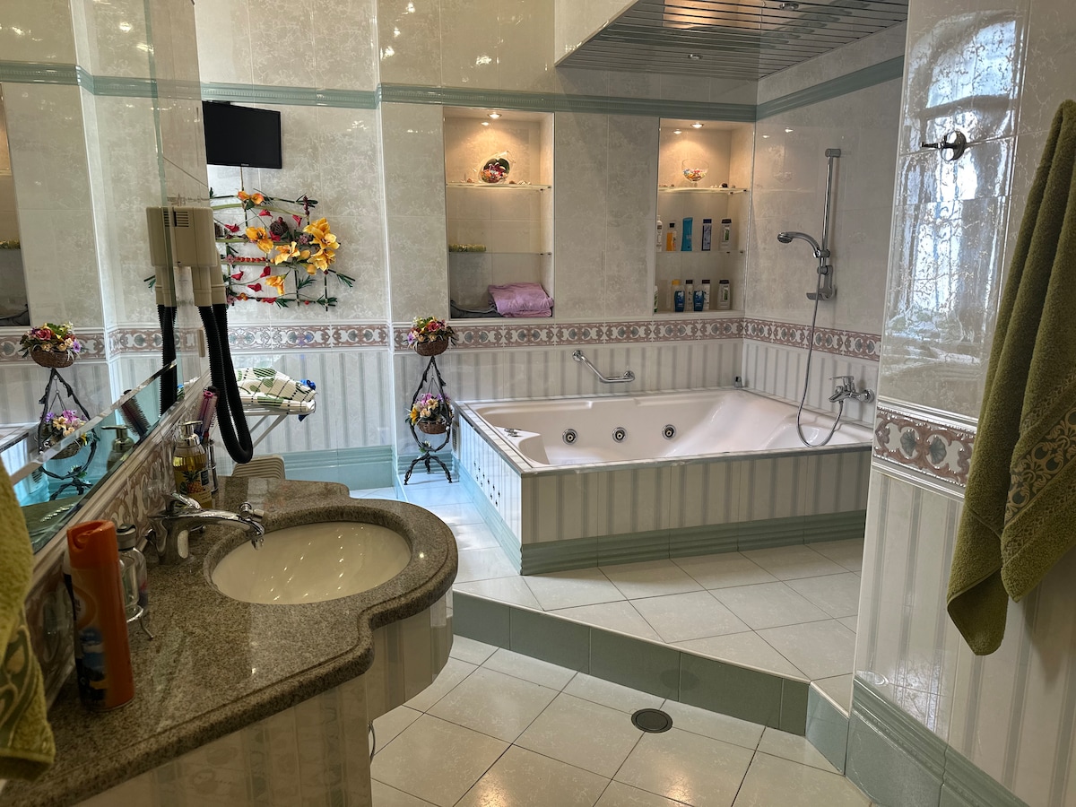 A spacious bathroom features a large whirlpool tub, surrounded by tiled walls and decorative accents. A double sink with a granite countertop offers ample space. Shelves display various toiletries, while soft lighting enhances the inviting atmosphere.