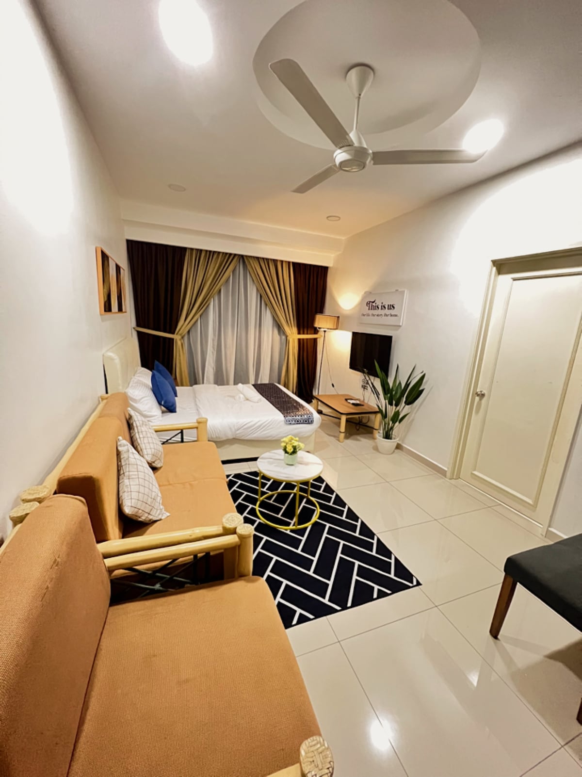 A cozy unit displays a spacious bed with white linens, complemented by plush pillows. A seating area features two wooden chairs and a light brown sofa, creating a relaxed vibe. A small round table sits centrally, while soft lighting and plants enhance the overall ambiance.