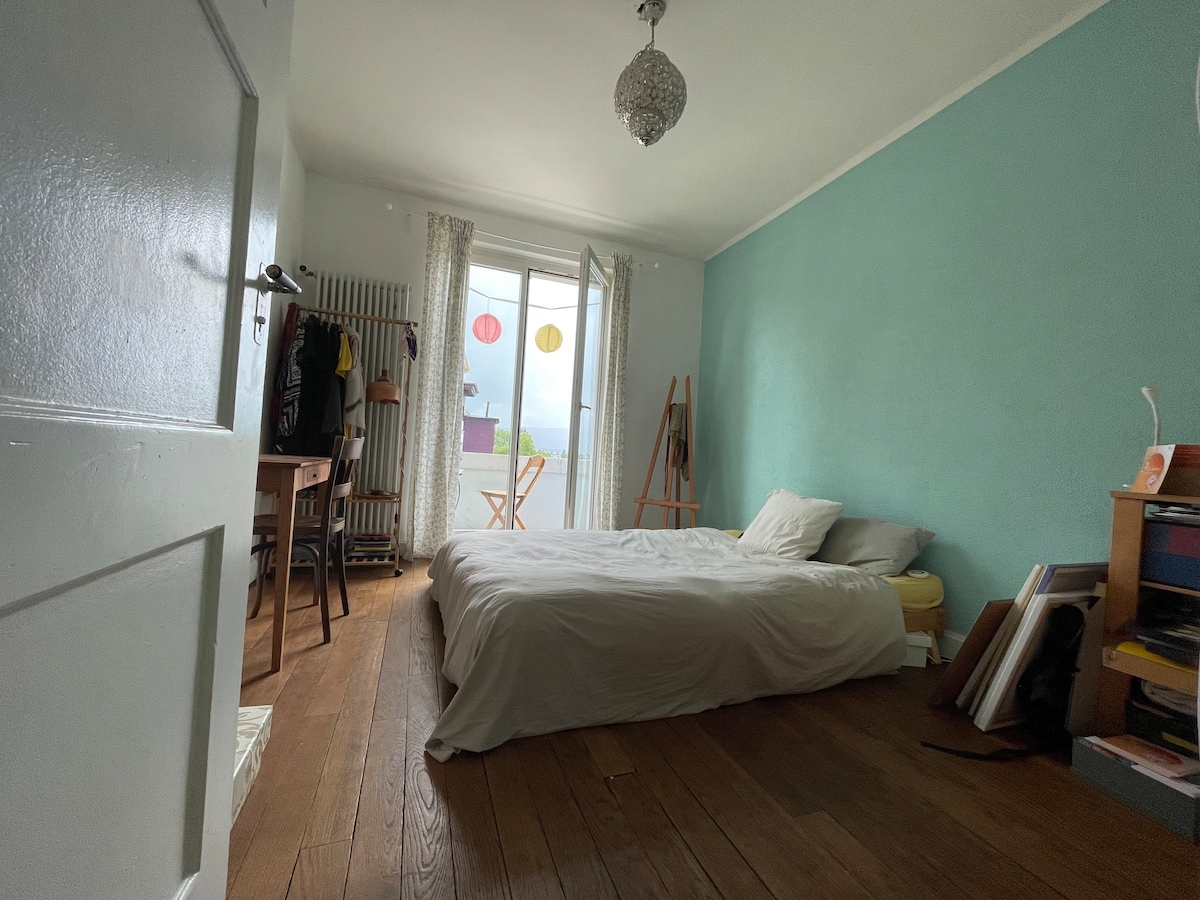 A bright bedroom features a spacious bed with neutral bedding and light wooden floors. A door opens to a balcony, while colorful lanterns can be seen through the curtains. A small table and easel are positioned near the window, enhancing the room's inviting feel.