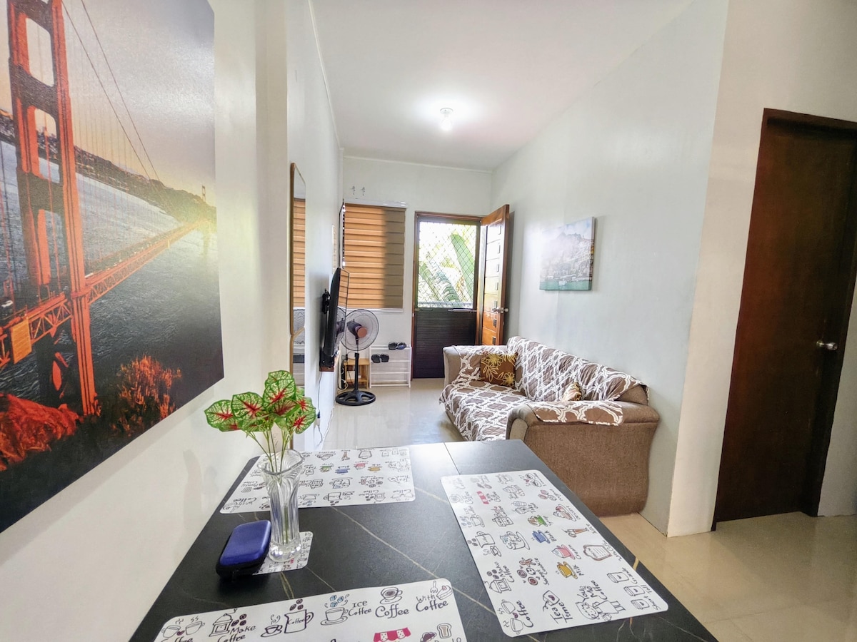 A welcoming living area features a comfortable sofa and a flat-screen TV against a bright wall. A small dining table with artistic placemats occupies the center. Natural light filters through the windows, and a decorative wall photo of a bridge enhances the space.