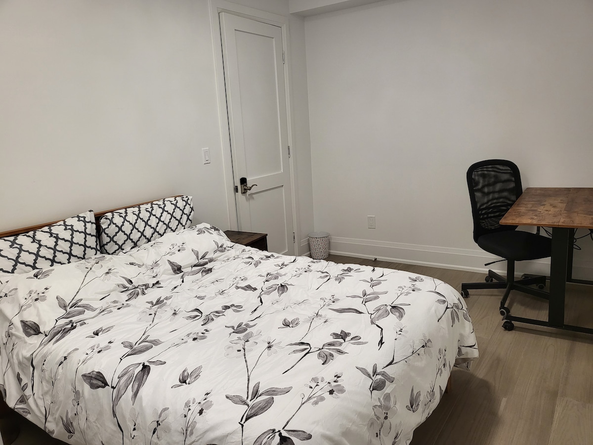 A spacious bedroom is furnished with a large bed featuring a floral-patterned duvet cover. A simple wooden desk and a black office chair are positioned nearby, and a wastebasket is placed in the corner. The room is well-lit with neutral-colored walls.