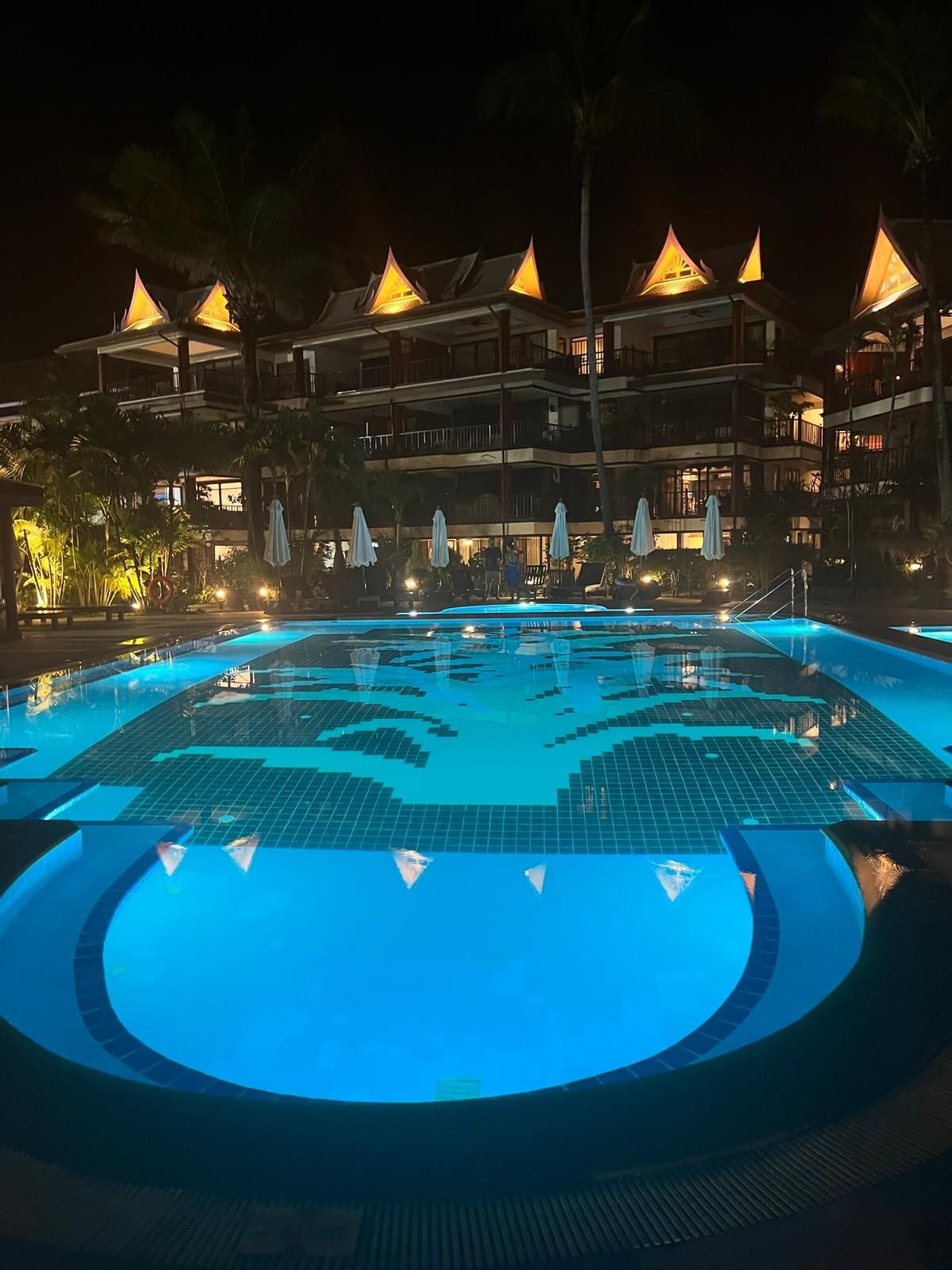 A spacious swimming pool is illuminated with soft blue lights, creating a serene ambiance. Surrounding the pool, palm trees and tropical landscaping are complemented by well-placed lighting, highlighting the architectural features of the building in the background.