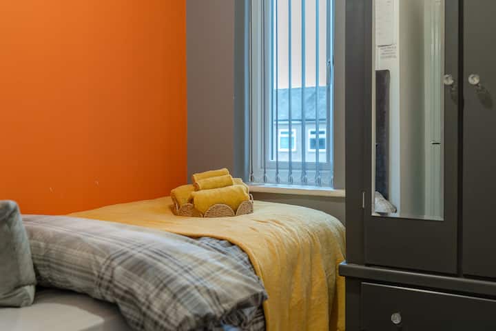 Kitanda kimoja kimewekwa karibu na dirisha na ukuta wa rangi ya machungwa.