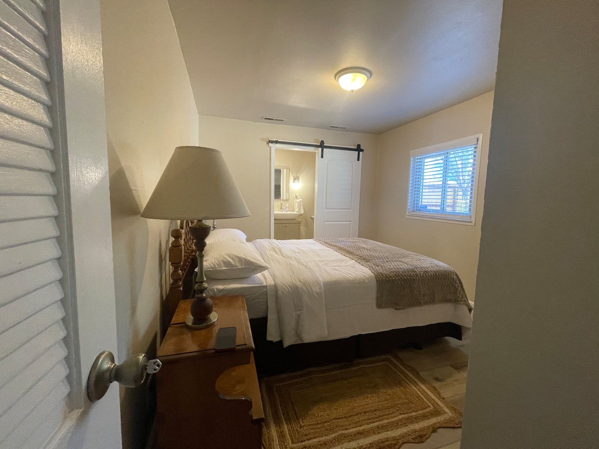 A calm bedroom features a spacious bed with white linens, complemented by a decorative throw. A bedside lamp is positioned on a wooden nightstand. Natural light enters through a window with blinds, illuminating the simple decor and adding warmth to the space.