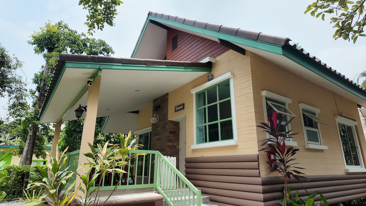 A charming exterior of a house is presented, featuring a sloped roof and a combination of vibrant green and neutral-toned walls. Lush greenery and colorful plants frame the entrance, which is enhanced by large windows allowing natural light to fill the space.