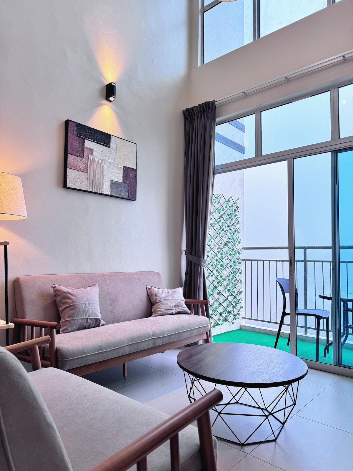 The living area features two modern sofas and a round, wireframe coffee table. Large glass doors offer access to a balcony with a view, while soft lighting enhances the space. A wall art piece adds a pop of color, complementing the neutral tones.