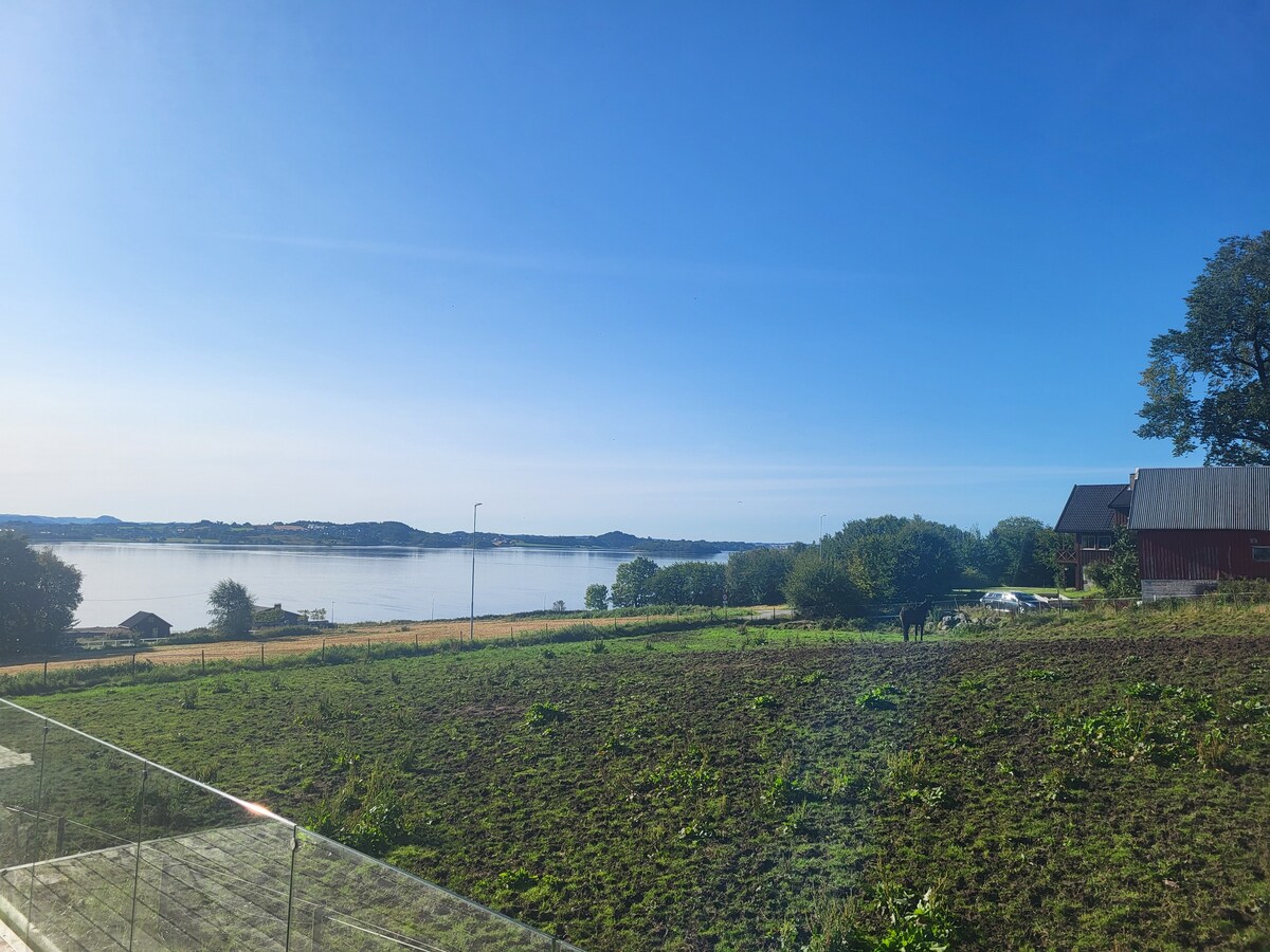 A panoramic view of Hafrsfjord showcases calm waters under a clear blue sky. Lush greenery in the foreground contrasts with the serene landscape, while distant hills provide depth. Nearby, a charming red building is visible, adding a touch of local architecture to the scene.
