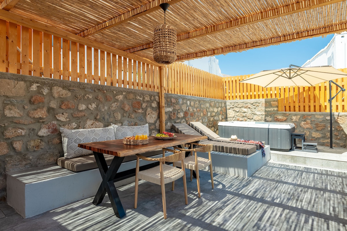 An outdoor terrace is presented, featuring a shaded dining area with a wooden table and light-colored chairs. Sun loungers are positioned nearby, and a jacuzzi is visible. The surrounding stone walls and bamboo overhead enhance the space, providing a relaxed atmosphere.