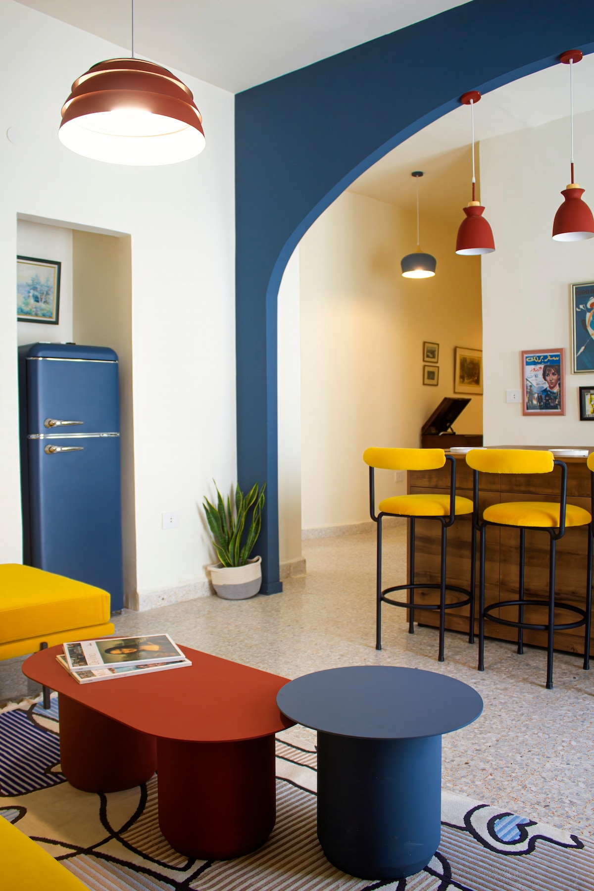A bright living area features a yellow seating area and a red and blue coffee table. Behind, a bar with high stools enhances the space. Soft lighting from overhead fixtures complements the blue accent wall and framed artwork, creating a cheerful atmosphere.