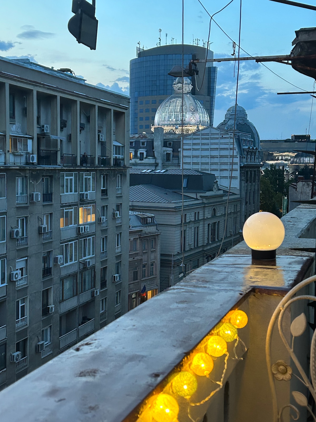 A balcony view reveals a city skyline at dusk, featuring a mix of modern and historical buildings. Soft lighting from a globe lamp and string lights adds a warm glow to the scene. The sky transitions from blue to hues of orange and pink.