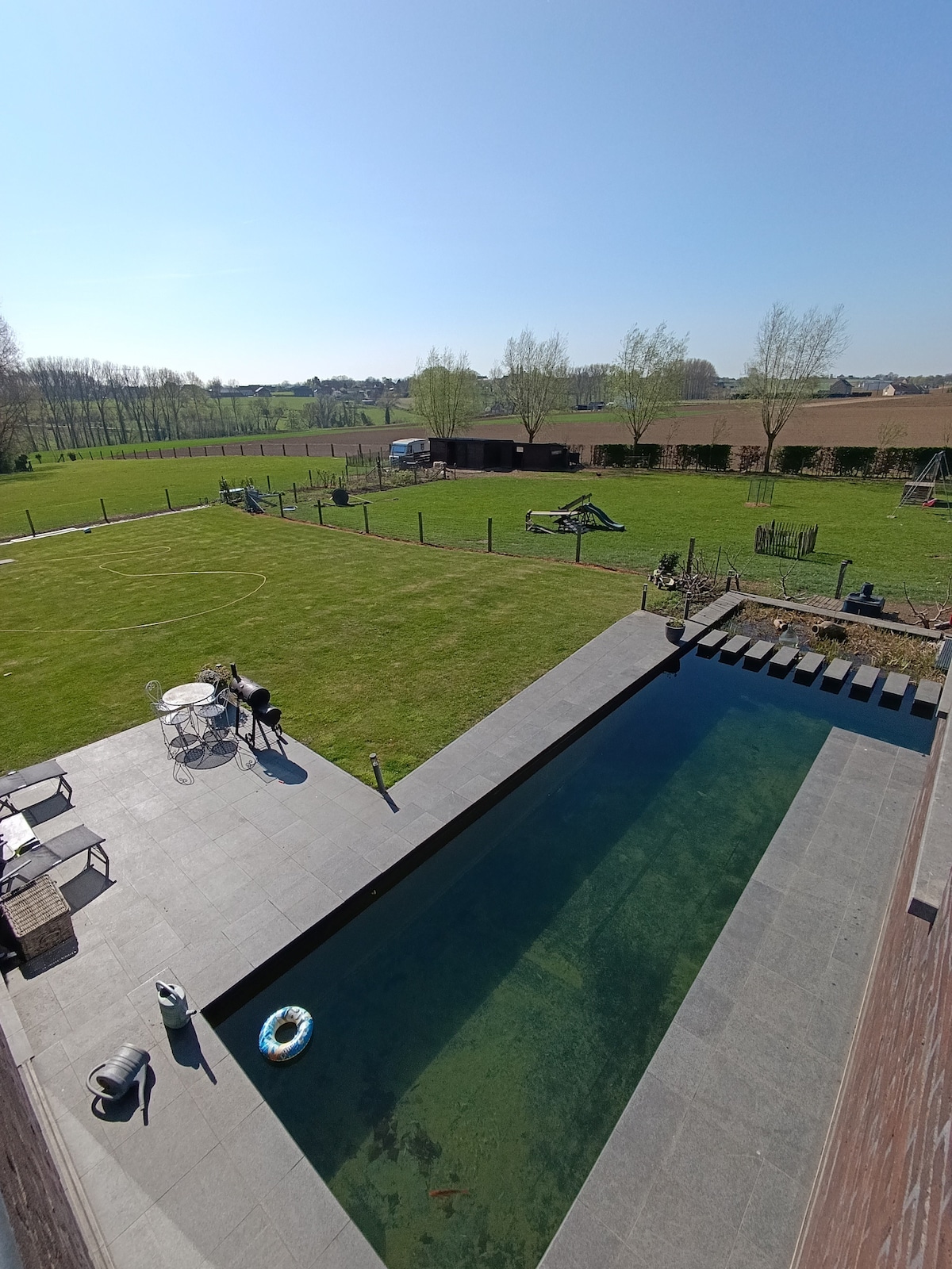 An aerial view reveals a spacious grassy area, outlined by trees and fencing. A tranquil swimming pond is centrally located, accompanied by a seating area with chairs and a table on the patio. Nearby, farm animals can be seen in their enclosures.