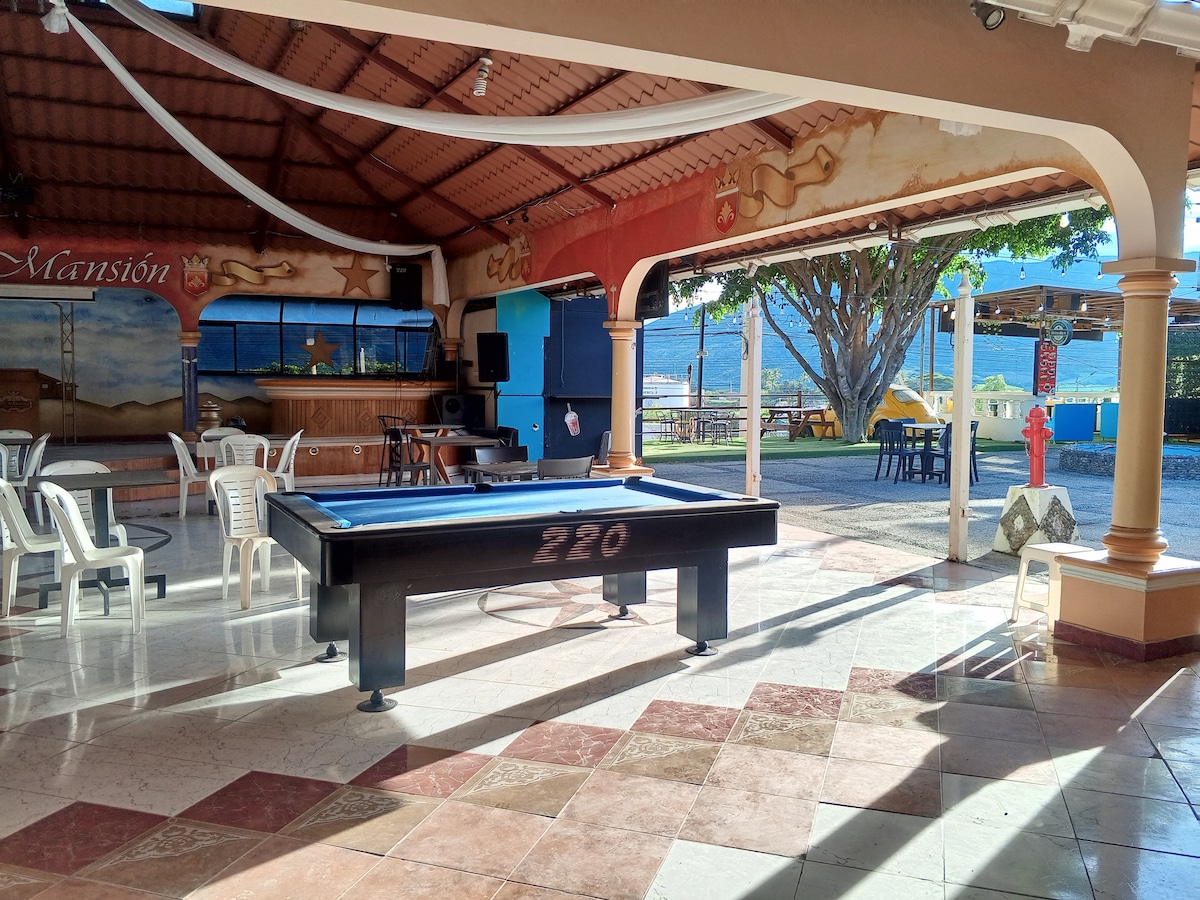 An open recreational area is featured, with a classic pool table at its center. Surrounding seating includes several white chairs. Large windows provide views of the outdoors, where trees and a scenic landscape are visible, creating a bright and inviting atmosphere.